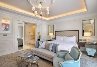 A hotel room with a large bed, a sofa, a small table, and a chair. There is a mirror and a desk with a lamp.