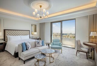 A spacious hotel room with a large bed, a sofa, two round tables, a chair, and a view of the city.