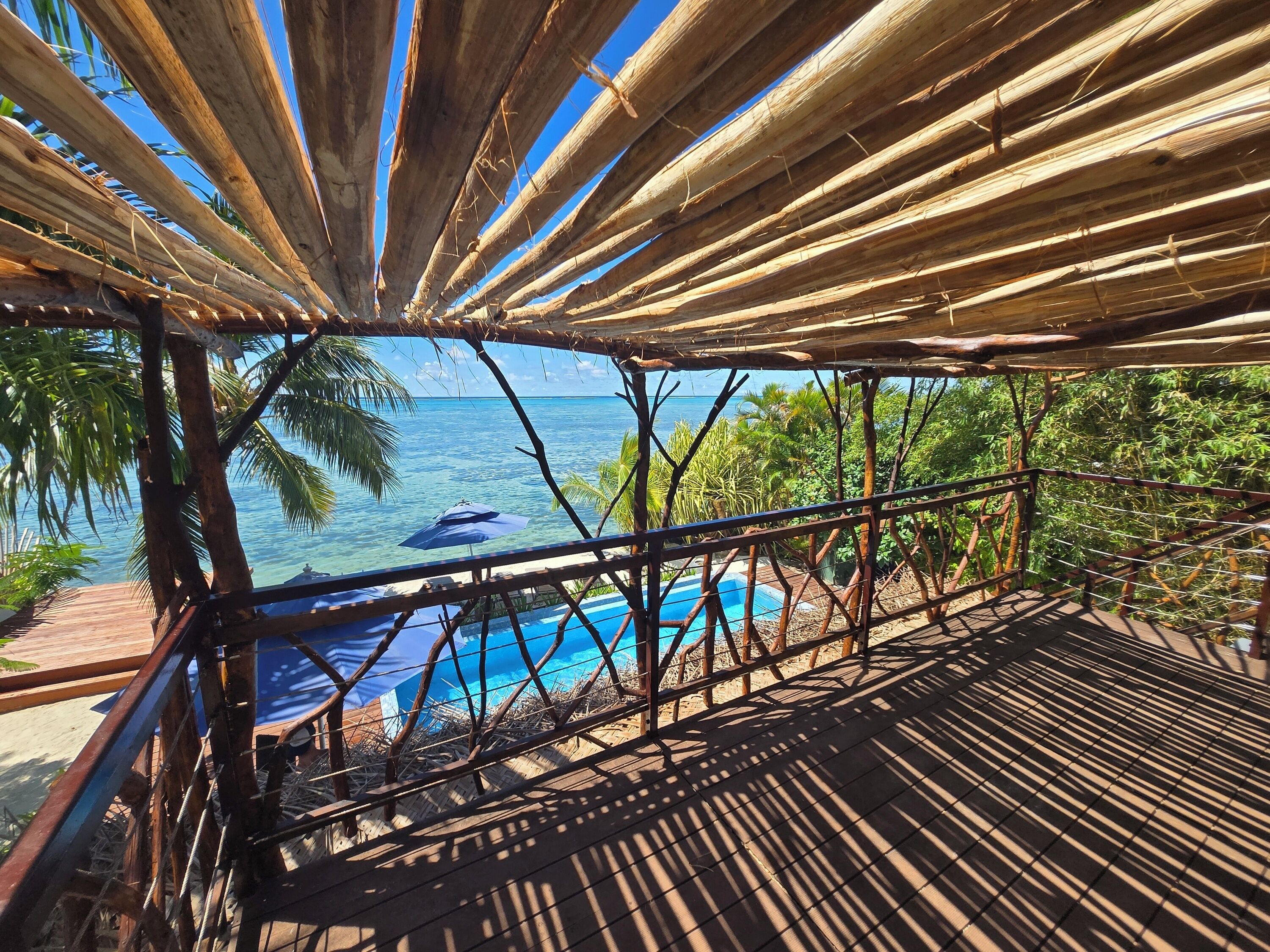 A wooden deck overlooking a pool with a view of the ocean and palm trees.