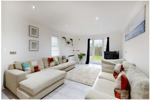 A spacious living room with a beige sofa set, a flat-screen TV, and a large window with curtains. - LEA - Elegant 1bed flat w/parking 5 min to Hackney Wick (London)