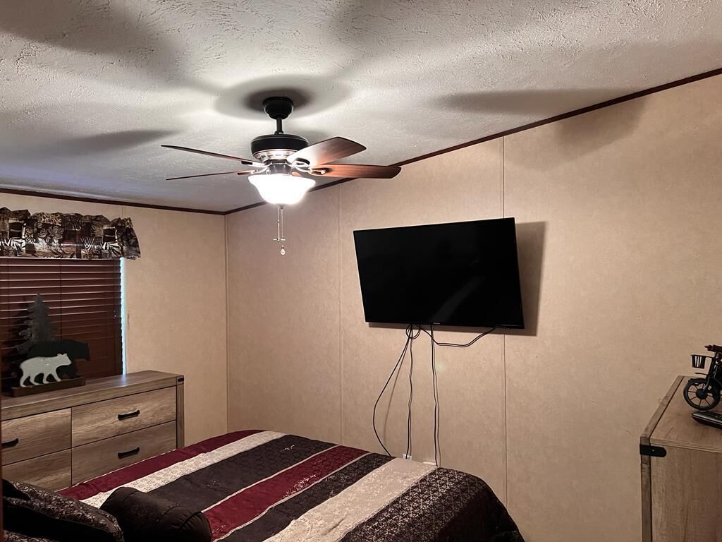 A bedroom with a bed, a ceiling fan, a mounted TV, a dresser, and a window with blinds.