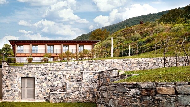 Un bâtiment moderne, avec un mur de pierre à l’avant, situé au pied d’une colline, sous un ciel dégagé.