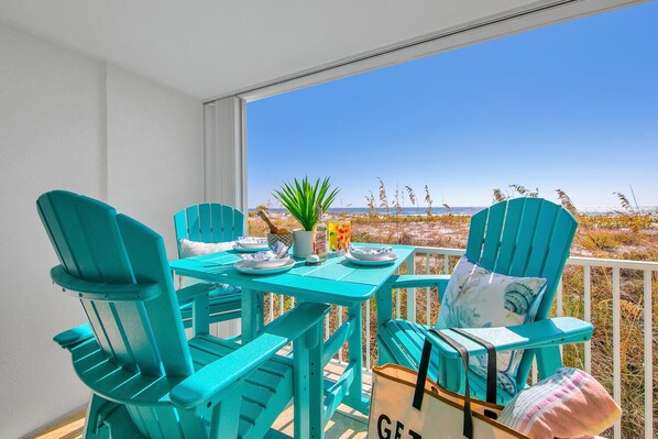 A balcony with turquoise chairs and a table, overlooking a beach with tall grasses and a clear blue sky.