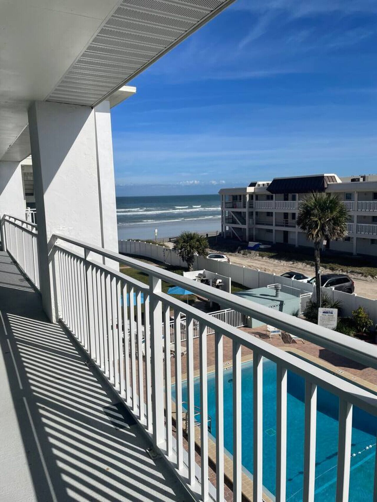 A balcony with a view of the ocean, a swimming pool, and a building with multiple floors.