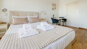 A neatly made bed with two towels, a bedside table with a lamp, a desk with a chair, and a door in a hotel room. - GuestReady - Port Studio 3 (Vila Nova de Gaia)