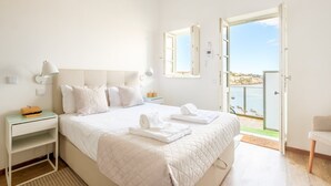A bedroom with a bed, bedside tables, a nightstand, a window with a view of the sea, and an open door leading outside. - GuestReady - Port Studio 4 (Vila Nova de Gaia)