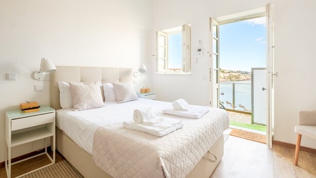 A bedroom with a bed, bedside tables, a nightstand, a window with a view of the sea, and an open door leading outside. - GuestReady - Port Studio 4 (Vila Nova de Gaia)
