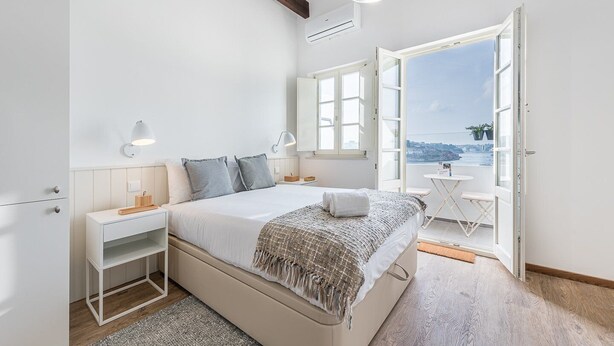 A bedroom with a bed, bedside tables, a nightstand, a window, and an open door leading to a balcony with a view. - GuestReady - Port Studio 1 (Vila Nova de Gaia)