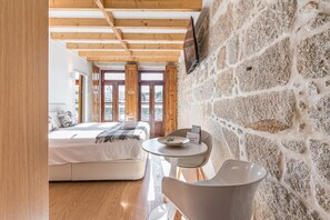 A bedroom with a bed, a round table, and a chair. There are wooden beams on the ceiling and a stone wall.