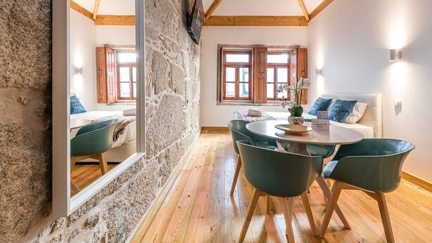 A cozy room with a stone wall, wooden beams, a round table with chairs, a bed with pillows, and a window with wooden shutters. - GuestReady - Miragaia Stone Studio 2T (Porto)