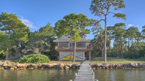 A two-story house with a balcony, a dock leading to a body of water, and surrounding trees. - Welcome to Bienvenue! (St. George Island)