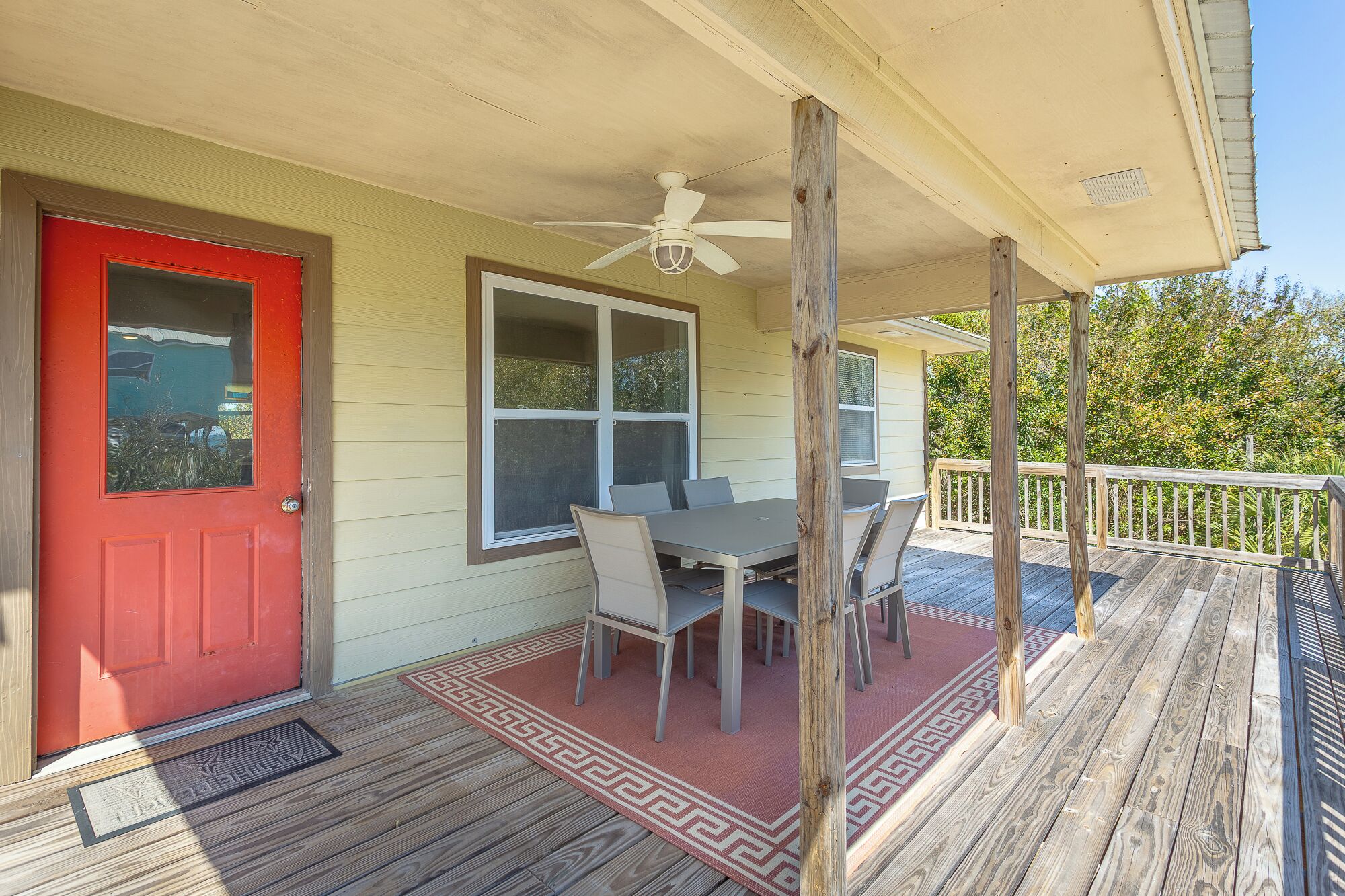 A covered porch with a dining table and chairs, a red door, and a ceiling fan.