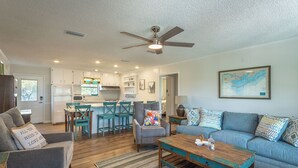A modern living room with a sofa, coffee table, and chairs. There is a kitchen area with a refrigerator, microwave, and cabinets. A ceiling fan is visible, and a map is displayed on the wall. - Easy Beach Access - Newly Updated! (St. George Island)