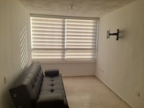 Una habitación con sofá, ventana con persianas, luz empotrada en la pared y suelo de baldosas.