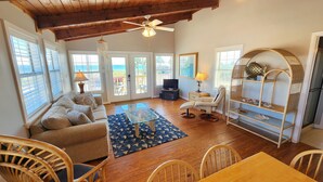 A spacious living room with a wooden ceiling, a glass coffee table, a sofa, a wicker chair, a dining table, and a wicker shelf. - Welcome to Catbird Seat! (St. George Island)