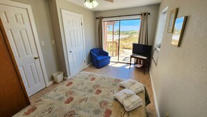 A bedroom with a bed, a blue chair, a television, and a view of the outdoors. - Welcome to Catbird Seat! (St. George Island)