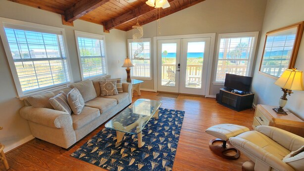 A spacious living room with a beige sofa, a glass coffee table, and a television. Large windows offer views of the outdoors. - Welcome to Catbird Seat! (St. George Island)