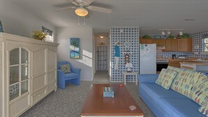 Un salon avec un canapé bleu, une table basse en bois, un fauteuil bleu, un meuble blanc et un coin cuisine doté de meubles en bois et d’un réfrigérateur.