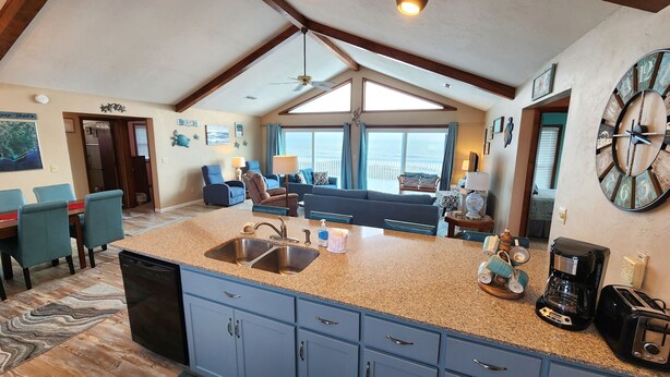 A spacious kitchen with granite countertops, a sink, and blue cabinets. There is a dining area with blue chairs and a table. The room has large windows and a ceiling fan. - Beachfront with large sundeck! (St. George Island)