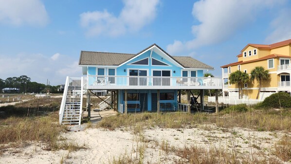 A two-story beach house with a balcony, a wooden deck, and a sandy beach in the background.