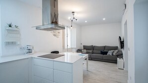 A modern kitchen with white cabinetry, a central island, and a built-in stove. There is a grey sofa in the living area with a coffee machine on the counter. - Apartment Las Lectoras de Cadiz (Cádiz)