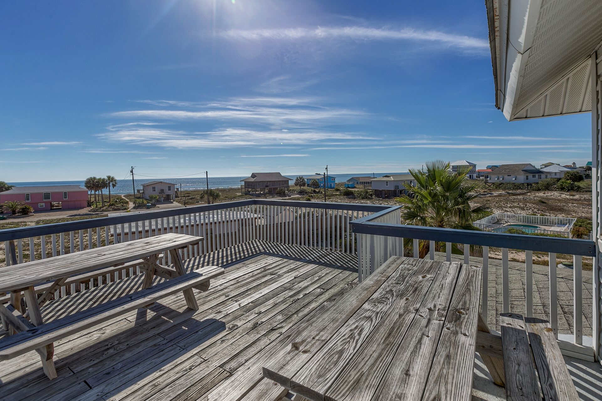Une terrasse en bois avec des tables de pique-nique, donnant sur une plage bordée de maisons, sous un ciel dégagé.