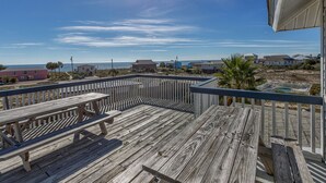 Une terrasse en bois avec des tables de pique-nique, donnant sur une plage bordée de maisons, sous un ciel dégagé.