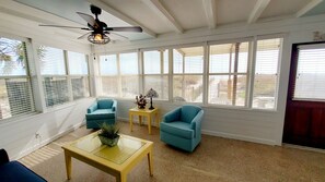 A sunlit room with a ceiling fan, two blue armchairs, a small table with a plant, and large windows with blinds.