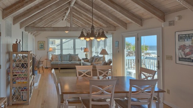 A spacious living area with a wooden dining table, white chairs, and a sofa. There is a bookshelf with books and decorative items. Large windows provide a view of the outdoors. - Beachfront! Large sundeck with screened in porch! (St. George Island)