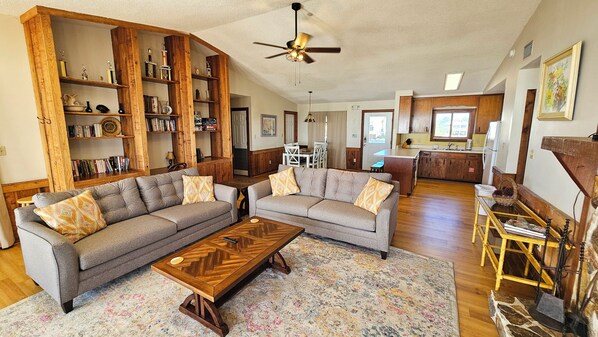 A spacious living room with two grey sofas, a wooden coffee table, and a large wooden bookshelf filled with trophies. - Prima Villa: 4 BR, 2 BA House in St. George Island, Sleeps 10 (St. George Island)