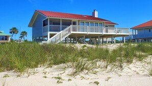 Uma casa à beira-mar com telhado vermelho, varanda coberta e uma praia arenosa em primeiro plano.