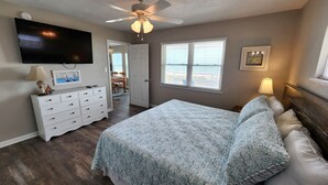 Un dormitorio con cama, cómoda, televisor, ventilador de techo y ventanas.