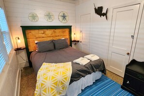 Une chambre avec une tête de lit en bois, un lit avec une literie grise, un coussin jaune et un tapis à rayures bleues. Les murs sont décorés et la porte est ornée d’un motif représentant un poisson.