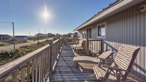 Una terraza de madera con mobiliario exterior, cielo despejado y una vista distante de casas.