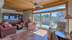 A spacious living room with a large TV, comfortable seating, a ceiling fan, and a sliding glass door leading to a patio. - BAY FRONT WITH PRIVATE DOCK! HOT TUB! (St. George Island)
