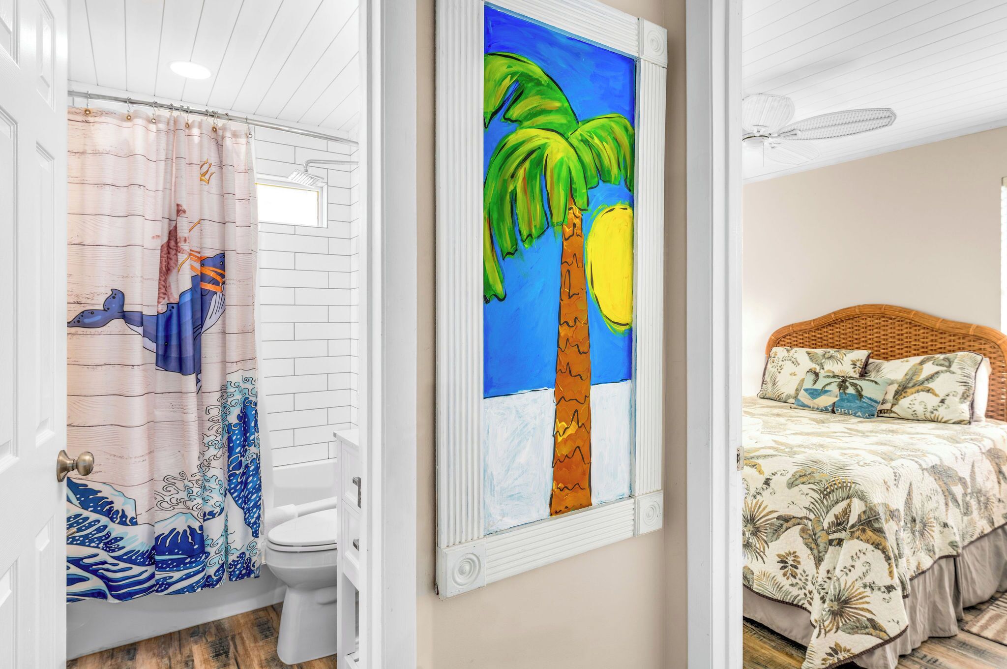 A bathroom with a shower curtain depicting a blue whale, a toilet, and a wall-mounted painting of a palm tree.