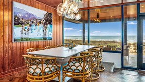 A dining area with a large table and wicker chairs, a wooden wall with a framed picture, and a view of the beach through large glass doors. - BEACHFRONT! PRIVATE POOL! All new renovation. (St. George Island)