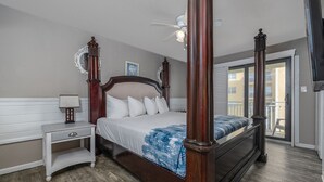 A four-poster bed with white bedding, a nightstand with a lamp, and a view of the outside balcony.