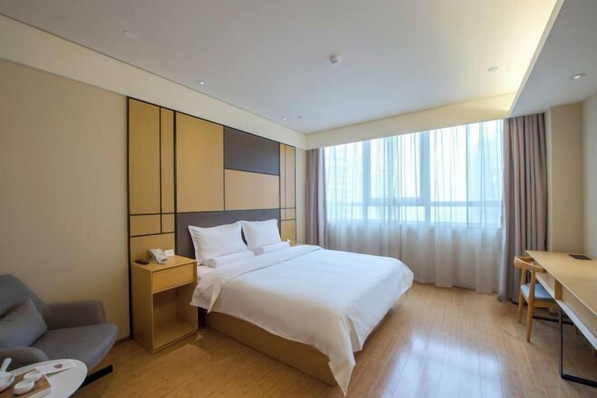 A hotel room with a bed, bedside table, chair, desk, and large window with curtains.