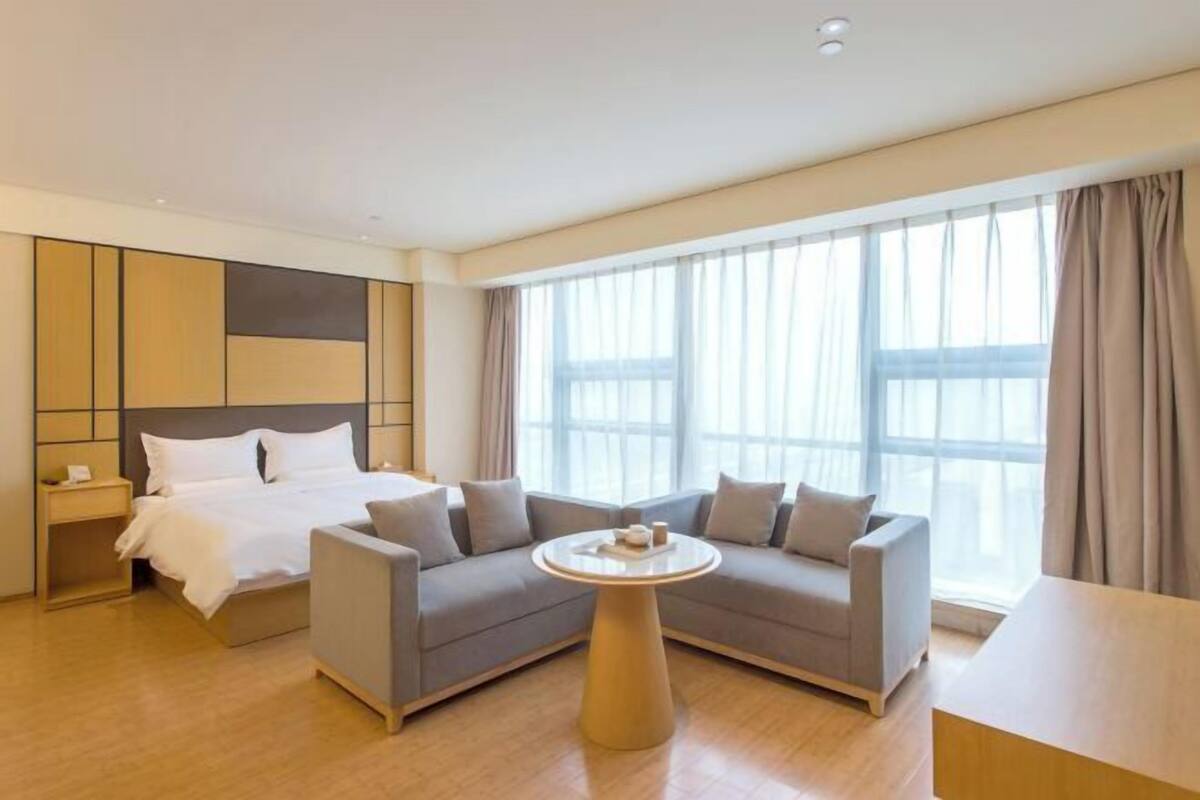 A modern hotel room with a bed, a gray sofa, a round coffee table, and large windows with curtains.