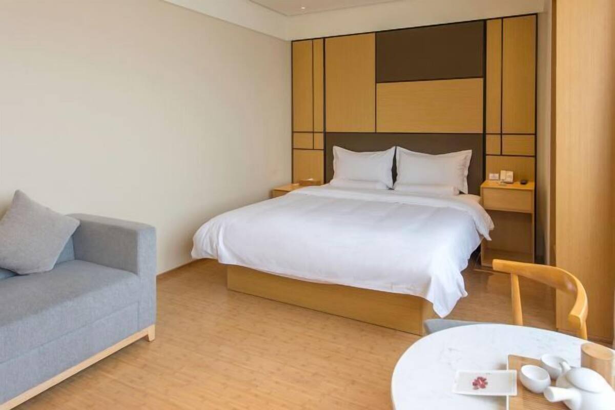 A hotel room with a bed, a grey sofa, a round table with a vase, and a wooden headboard.