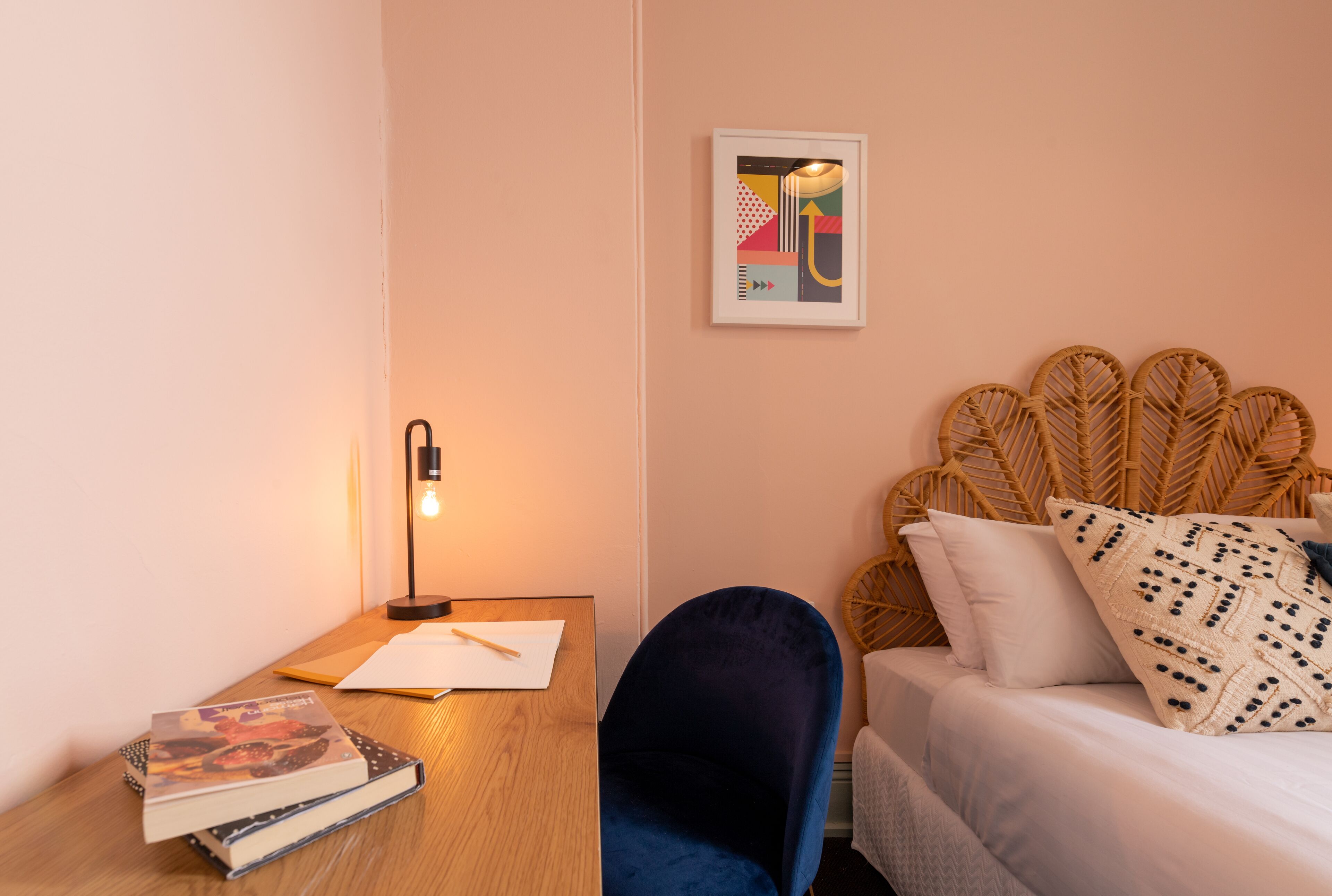 Un dormitorio con una cama, una mesita de noche con libros, una lámpara y una obra de arte en la pared.