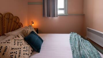 A bedroom with a bed, pillows, a bedside lamp, a window with curtains, and a wall-mounted heater.