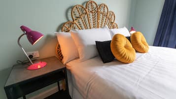 Una cama bien hecha con sábanas blancas, un cabecero de ratán y dos almohadas color mostaza. Una mesita de noche con una lámpara rosa y un tapete para ratón está al lado de la cama.