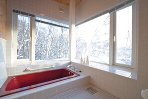 Un baño con una bañera roja, ventanas amplias y vistas a árboles nevados.