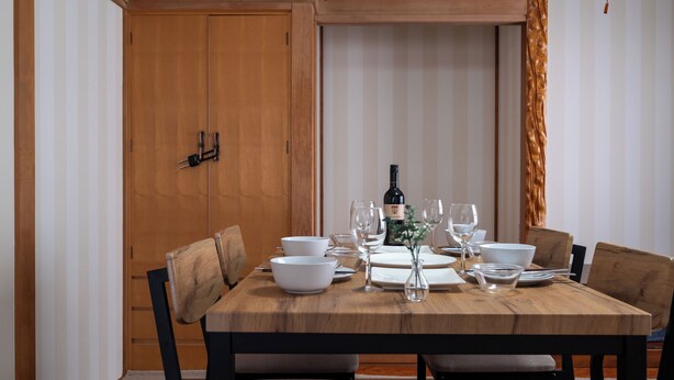 Una mesa de comedor con vajilla blanca, copas de vino y una botella, rodeada de sillas.