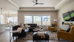 A spacious living room with a black leather sofa, a yellow armchair, and a coffee table. There is a ceiling fan, a chandelier, and a large window with a view of the backyard.