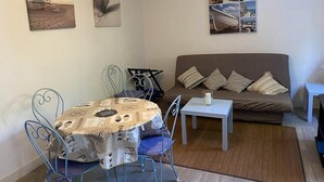 Una sala de estar acogedora con un sofá marrón, una mesita blanca pequeña y una mesa de comedor con sillas metálicas. Hay tres obras de arte enmarcadas en las paredes.
