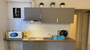 Una cocina compacta con armarios grises, un microondas, una cafetera y un lavamanos. Hay tres plantas en macetas sobre la encimera y un cubo de basura a la izquierda.
