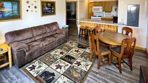 Una sala de estar acogedora con un sofá marrón, una mesa de comedor de madera con sillas, un tapete estampado y un ventilador de techo.